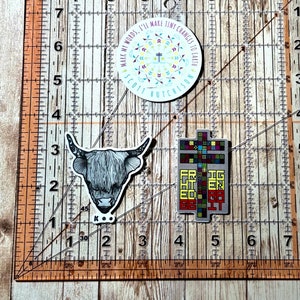 May include: Three stickers on a white surface with a ruler. The first sticker is a round sticker with a colorful design and the text "Mark my words, I'll make tiny changes to earth. Scott Hutchison". The second sticker is a black and white illustration of a cow's head. The third sticker is a colorful geometric design with the text "For the Greater Good".