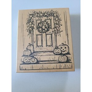 May include: A wooden stamp featuring a detailed illustration of a front door decorated for autumn. The door is adorned with a wreath and garland, with pumpkins of various sizes on the steps. The design is in black ink.