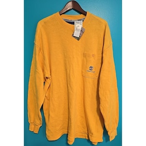 May include: Mustard yellow long-sleeve shirt with a pocket on the left chest. The shirt has a Timberland logo embroidered on the pocket. A price tag is attached to the collar. The shirt is hanging on a wooden hanger.
