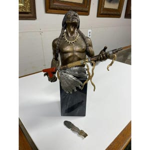 May include: Bronze bust of a Native American man with long braids, wearing a bone necklace, and holding a spear with feathers. The bust is mounted on a black marble base. A detached feather lies nearby.