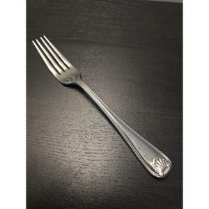 May include: A silver-toned metal dinner fork featuring four tines and a slender handle. The handle is decorated with a raised shell motif at the base and subtle vertical line detailing that runs along the length of the stem toward the fork head.