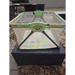 May include: A vintage Cabbage Patch Kids playard. The playard has a green frame with a mesh interior. The top edge features a colorful pattern. The Cabbage Patch Kids logo is visible on the front. The playard is set on a black surface.