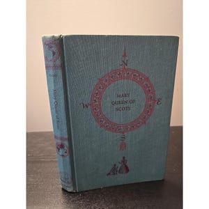 May include: A teal hardcover book titled Mary Queen of Scots. The front cover features a red compass rose design with the text Mary Queen of Scots in the center and a small illustration of two figures at the bottom. The spine shows text.