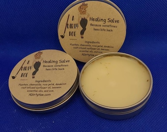 Op de afbeelding: Twee metalen blikken met helende zalf met een bruin etiket waarop staat "A Dirty Hoe Healing Salve Because sometimes hoes bite back" en de ingrediënten worden vermeld: weegbree, kamille, rozenblaadjes, paardenbloemwortel, zonnebloemolie, bijenwas, etherische oliën en liefde. Het websiteadres ADirtyHoe.com staat op het etiket gedrukt.