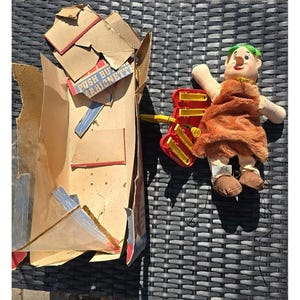 May include: A vintage marionette doll with a brown fur-like outfit, green hair, and a red and yellow control mechanism. The doll is next to its worn cardboard box, which has the text "PUSH BUY MARIONETTE".