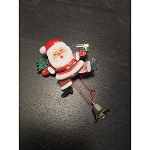 May include: A Christmas ornament featuring Santa Claus with a red hat and suit, holding a small green Christmas tree and a gift. A small gold bell hangs from a red string attached to the ornament.
