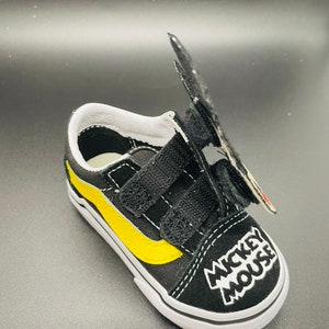 Puede incluir: Zapatillas Vans negras y amarillas con un dise&ntilde;o de Mickey Mouse y cierres de velcro.