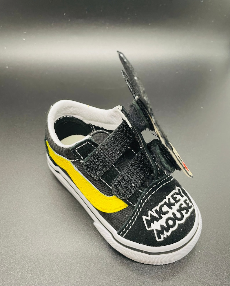 Puede incluir: Zapatillas Vans negras y amarillas con un dise&ntilde;o de Mickey Mouse y cierres de velcro.