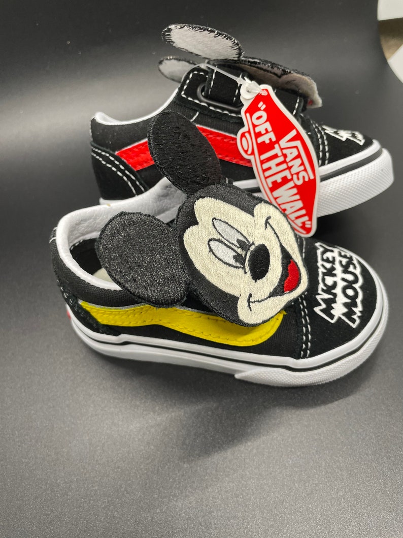 Puede incluir: Zapatillas Vans negras y amarillas con un personaje de Mickey Mouse en el lateral. Las zapatillas tienen una raya roja y una suela blanca. La etiqueta dice "Vans "Off The Wall"".