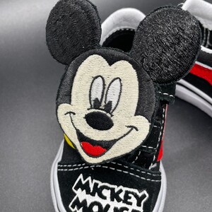 Puede incluir: Zapatillas Vans negras con un personaje de Mickey Mouse bordado en el lateral. El personaje tiene la boca roja, la nariz amarilla y los ojos blancos. Las palabras "Mickey Mouse" est&aacute;n bordadas en la punta del zapato.