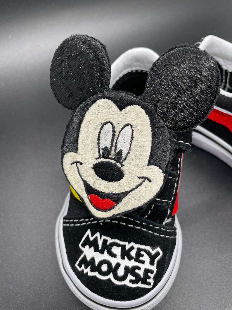 Puede incluir: Zapatillas Vans negras con un personaje de Mickey Mouse bordado en el lateral. El personaje tiene la boca roja, la nariz amarilla y los ojos blancos. Las palabras "Mickey Mouse" est&aacute;n bordadas en la punta del zapato.
