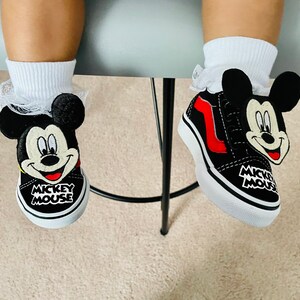 Puede incluir: Un par de zapatos Vans negros y rojos de Mickey Mouse con cordones blancos. Los zapatos tienen un dise&ntilde;o de cabeza de Mickey Mouse en blanco y negro en el lateral y las palabras "Mickey Mouse" en la parte posterior.