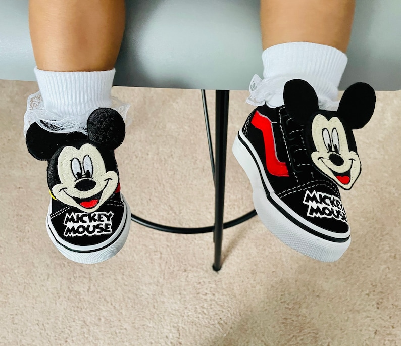 Puede incluir: Un par de zapatos Vans negros y rojos de Mickey Mouse con cordones blancos. Los zapatos tienen un dise&ntilde;o de cabeza de Mickey Mouse en blanco y negro en el lateral y las palabras "Mickey Mouse" en la parte posterior.