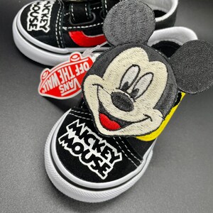 Puede incluir: Zapatillas Vans negras con dise&ntilde;o de Mickey Mouse. Las zapatillas tienen una parte superior de lona negra con una suela blanca. El dise&ntilde;o de Mickey Mouse est&aacute; bordado en el lateral de la zapatilla y presenta un contorno amarillo y mejillas rojas. Las zapatillas tambi&eacute;n tienen una correa negra con cierre de velcro.