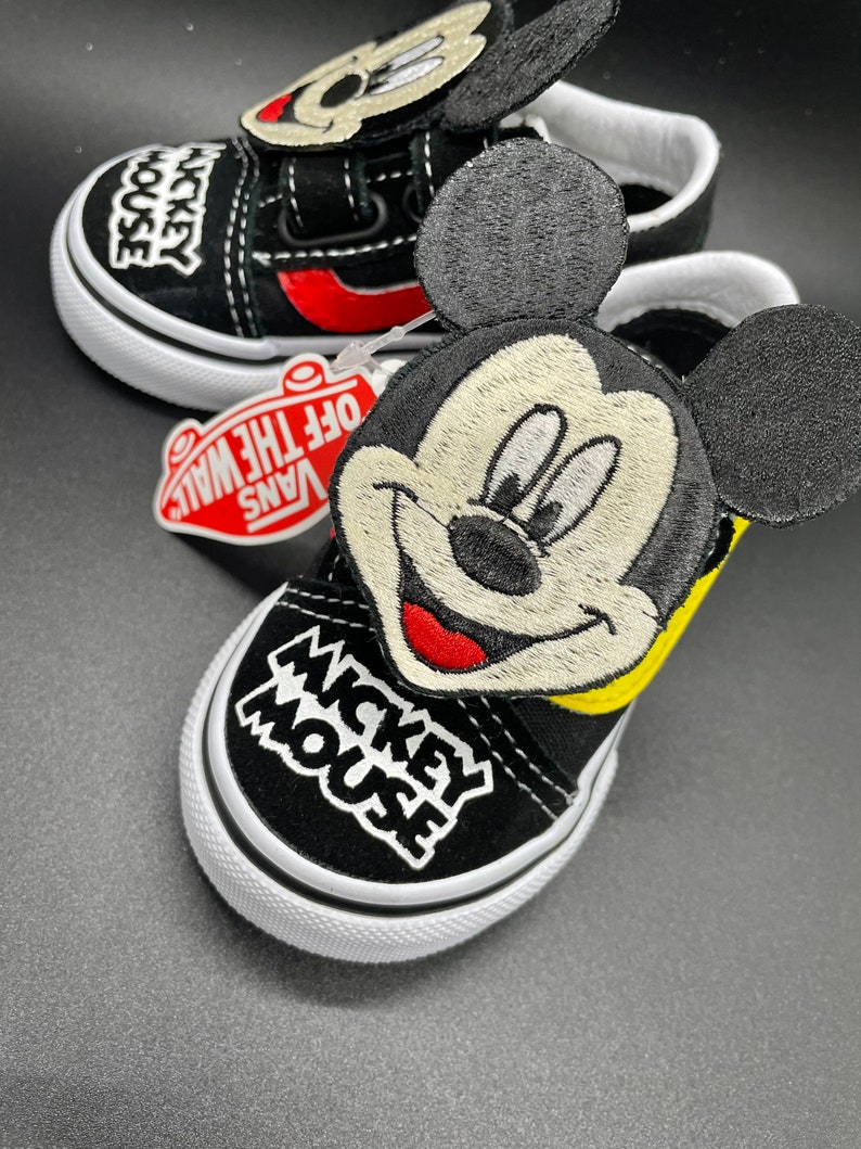 Puede incluir: Zapatillas Vans negras con dise&ntilde;o de Mickey Mouse. Las zapatillas tienen una parte superior de lona negra con una suela blanca. El dise&ntilde;o de Mickey Mouse est&aacute; bordado en el lateral de la zapatilla y presenta un contorno amarillo y mejillas rojas. Las zapatillas tambi&eacute;n tienen una correa negra con cierre de velcro.