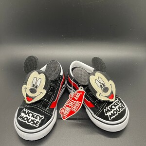 Puede incluir: Un par de zapatos Vans negros con rayas rojas y un personaje de Mickey Mouse en la parte delantera. Los zapatos tienen una suela blanca y el texto "Mickey Mouse" est&aacute; bordado en el lateral. Los zapatos vienen con una etiqueta de Vans que dice "Off The Wall."