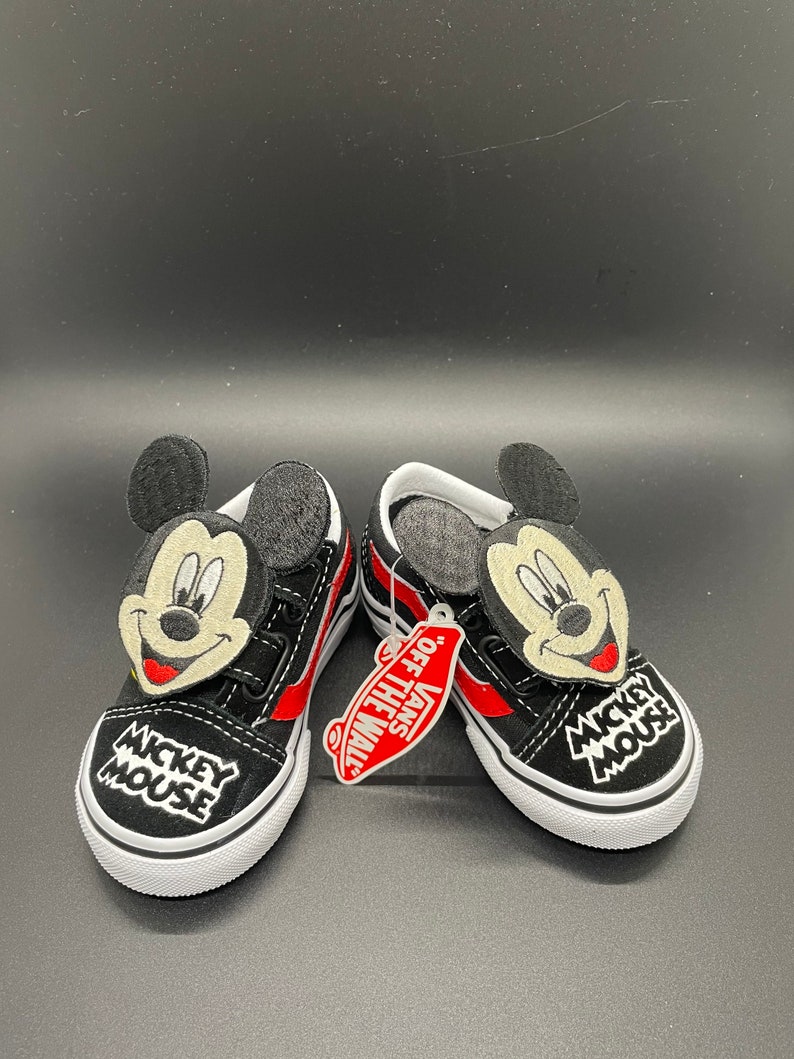 Puede incluir: Un par de zapatos Vans negros con rayas rojas y un personaje de Mickey Mouse en la parte delantera. Los zapatos tienen una suela blanca y el texto "Mickey Mouse" est&aacute; bordado en el lateral. Los zapatos vienen con una etiqueta de Vans que dice "Off The Wall."