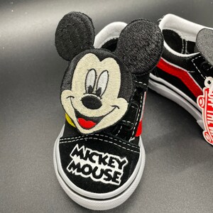 Puede incluir: Zapatillas Vans negras con una raya roja y un personaje de Mickey Mouse bordado en el lateral. Las zapatillas dicen "Mickey Mouse" en la parte delantera.