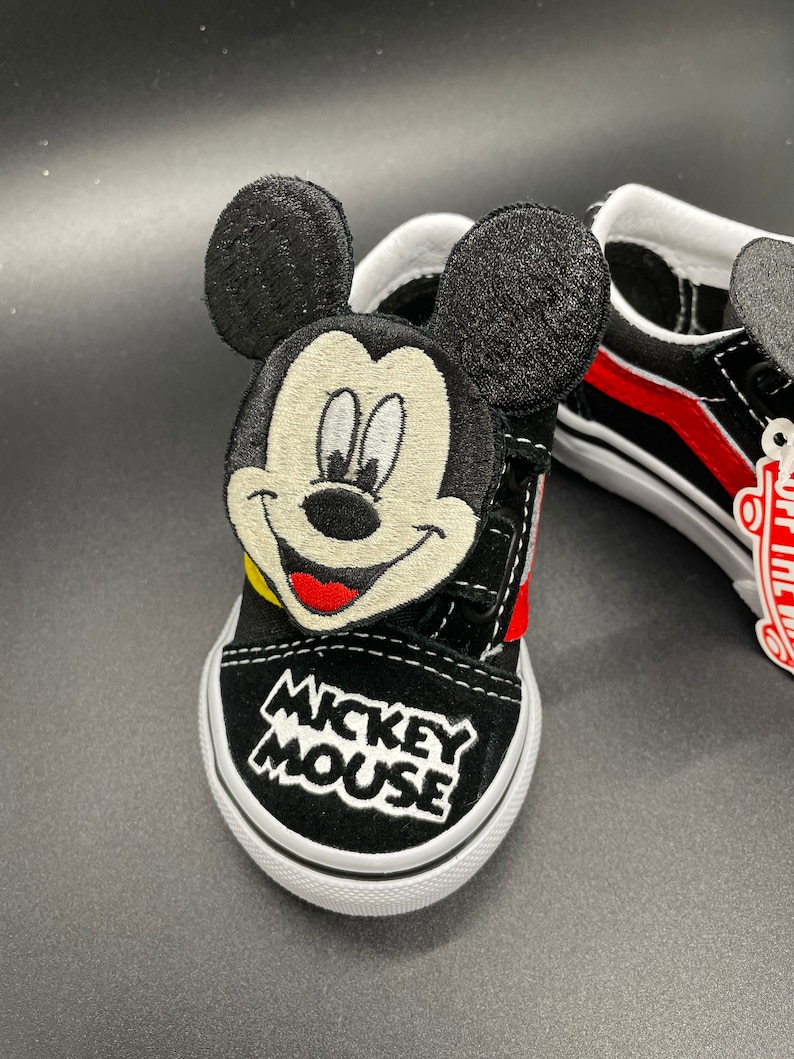 Puede incluir: Zapatillas Vans negras con una raya roja y un personaje de Mickey Mouse bordado en el lateral. Las zapatillas dicen "Mickey Mouse" en la parte delantera.