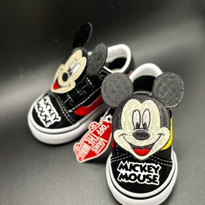Puede incluir: Zapatillas Vans negras y amarillas con un dise&ntilde;o de Mickey Mouse. Las zapatillas tienen una parte superior de lona negra con una raya roja y una suela blanca. El dise&ntilde;o de Mickey Mouse est&aacute; bordado en la parte delantera de las zapatillas e incluye la cara de Mickey, las orejas y las palabras "Mickey Mouse".
