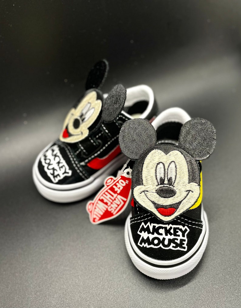 Puede incluir: Zapatillas Vans negras y amarillas con un dise&ntilde;o de Mickey Mouse. Las zapatillas tienen una parte superior de lona negra con una raya roja y una suela blanca. El dise&ntilde;o de Mickey Mouse est&aacute; bordado en la parte delantera de las zapatillas e incluye la cara de Mickey, las orejas y las palabras "Mickey Mouse".