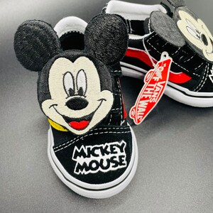 Puede incluir: Zapatillas Vans negras con un dise&ntilde;o de Mickey Mouse. Las zapatillas tienen detalles rojos y blancos y una cara de Mickey Mouse en blanco y negro en la parte delantera. Las zapatillas tambi&eacute;n tienen una etiqueta que dice "Vans Off The Wall."