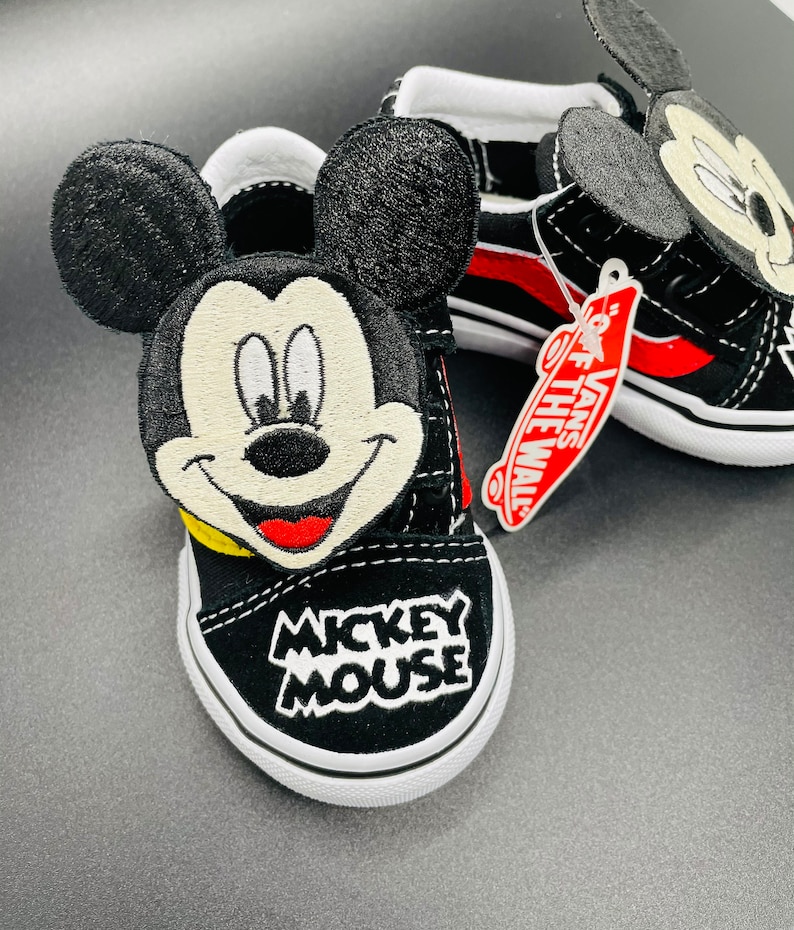 Puede incluir: Zapatillas Vans negras con un dise&ntilde;o de Mickey Mouse. Las zapatillas tienen detalles rojos y blancos y una cara de Mickey Mouse en blanco y negro en la parte delantera. Las zapatillas tambi&eacute;n tienen una etiqueta que dice "Vans Off The Wall."
