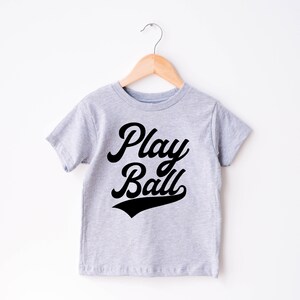 May include: A gray t-shirt with short sleeves and a black graphic that says "Play Ball" in a retro font.