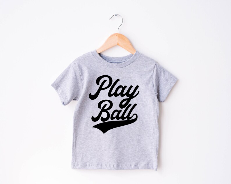 May include: A gray t-shirt with short sleeves and a black graphic that says "Play Ball" in a retro font.