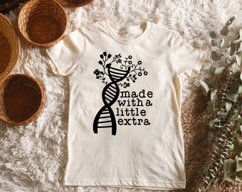 May include: A white t-shirt featuring a black graphic of a DNA strand with flowers growing from it. The text "made with a little extra" is below the graphic.