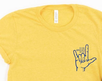 May include: Yellow t-shirt with a blue graphic of a hand making the American Sign Language sign for "I love you". The graphic also includes the text "Be Kind, Advocate, Include".