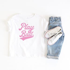 May include: White t-shirt with pink lettering that says "Play Ball" in a retro font style. The shirt is paired with blue denim jeans and white sneakers.