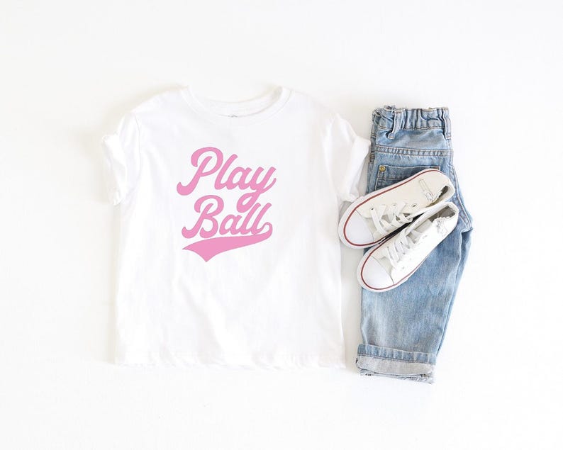 May include: White t-shirt with pink lettering that says "Play Ball" in a retro font style. The shirt is paired with blue denim jeans and white sneakers.