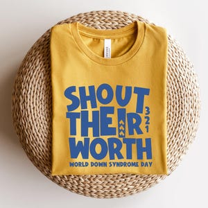May include: Mustard yellow t-shirt with the text "SHOUT THEIR WORTH" in blue, along with "WORLD DOWN SYNDROME DAY". The shirt is displayed on a woven, circular surface. The shirt is a crew neck style.