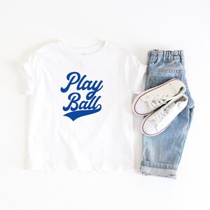 May include: White t-shirt with blue text that says "Play Ball" in a retro baseball font.
