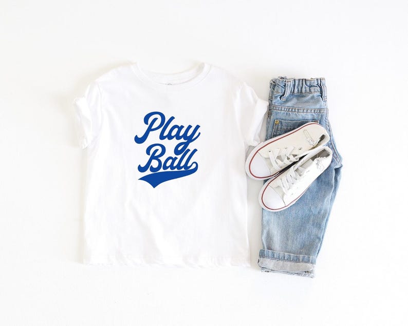 May include: White t-shirt with blue text that says "Play Ball" in a retro baseball font.
