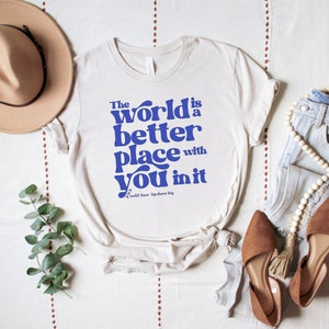 May include: A white t-shirt with blue text that reads "The world is a better place with you in it". The text is in a retro font style. The shirt is laid flat on a white surface with a brown hat, a pair of brown shoes, and a pair of blue jeans with a white beaded necklace.