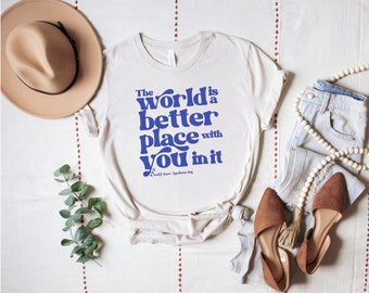 Puede incluir: Una camiseta blanca con texto azul que dice "The world is a better place with you in it". El texto está en un estilo de fuente retro. La camiseta está colocada sobre una superficie blanca con un sombrero marrón, un par de zapatos marrones y unos vaqueros azules con un collar de cuentas blancas.