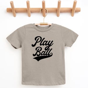 May include: A light gray t-shirt with short sleeves and a black graphic that says "Play Ball" in a retro font.