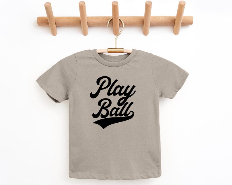 May include: A light gray t-shirt with short sleeves and a black graphic that says "Play Ball" in a retro font.
