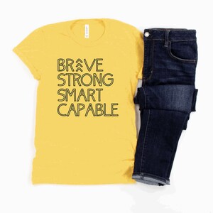 May include: A yellow t-shirt with black text that reads "BRAVE STRONG SMART CAPABLE". The shirt is paired with a pair of blue jeans.