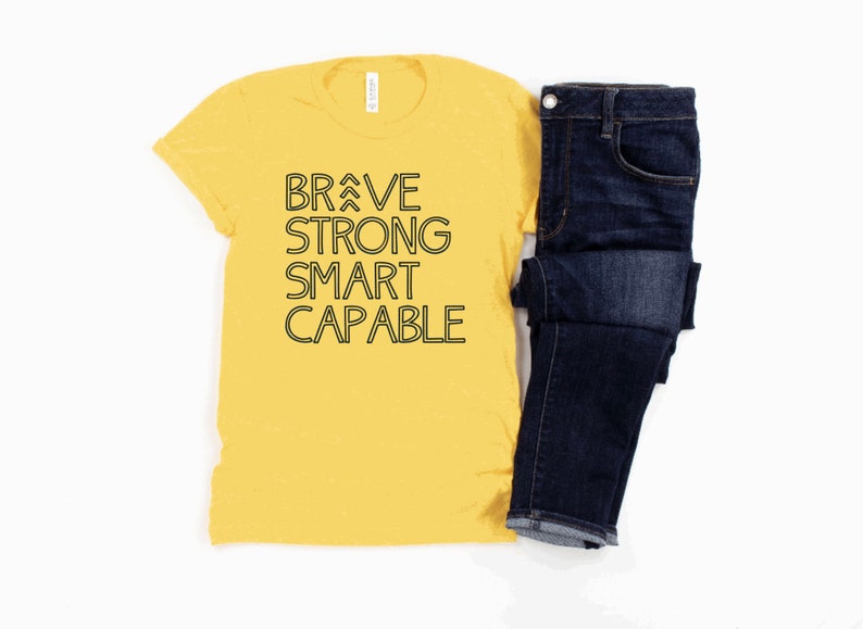 May include: A yellow t-shirt with black text that reads "BRAVE STRONG SMART CAPABLE". The shirt is paired with a pair of blue jeans.
