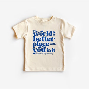 May include: A cream colored t-shirt with a blue graphic that reads "The world is a better place with you in it."  The graphic is in a retro font style.  The text "World Down Syndrome Day" is also included in smaller font below the graphic.