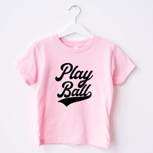 May include: Pink t-shirt with black text that says "Play Ball" in a retro baseball font.