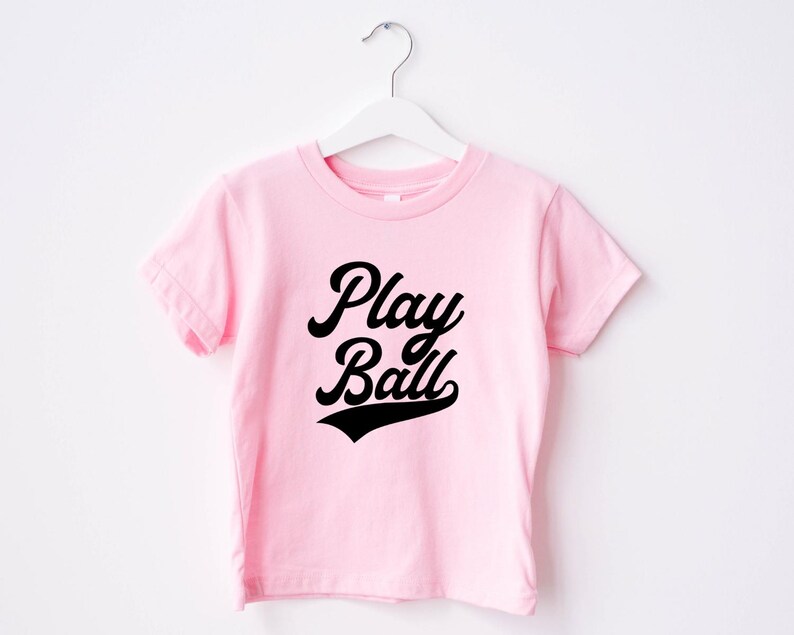 May include: Pink t-shirt with black text that says "Play Ball" in a retro baseball font.