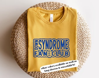 May include: Yellow t-shirt with blue text that reads "Down Syndrome Fan Club". The text is in a bold, blocky font. The shirt is on a woven straw surface. The text "other colours available as well as long sleeves & sweatshirts!" is printed on a white tag.