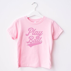 May include: Pink t-shirt with a pink "Play Ball" graphic in a retro font.