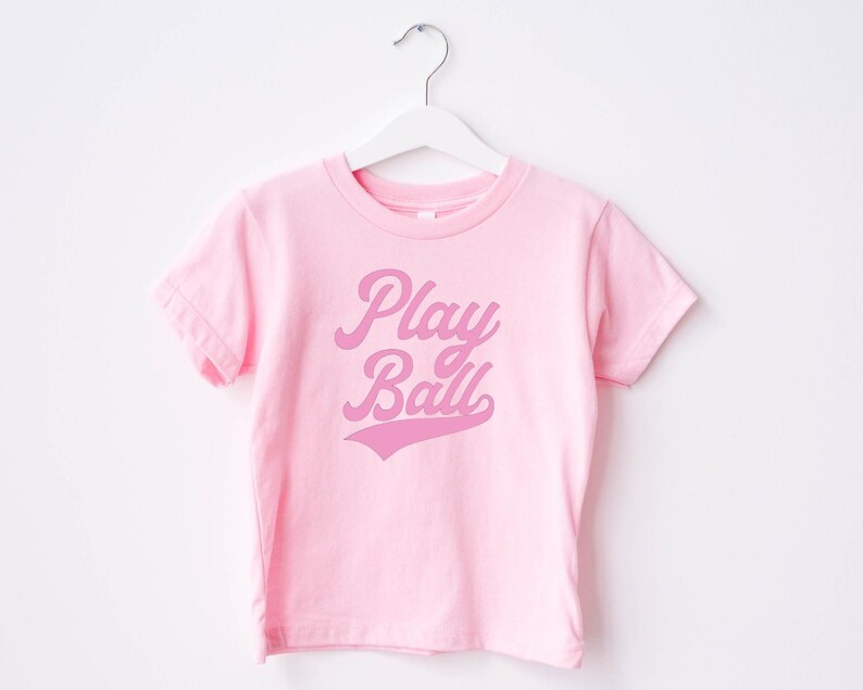 May include: Pink t-shirt with a pink "Play Ball" graphic in a retro font.