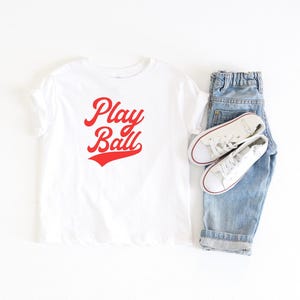 May include: White t-shirt with red text that says "Play Ball" in a retro font. The shirt is paired with blue jeans and white sneakers.