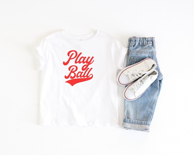 May include: White t-shirt with red text that says "Play Ball" in a retro font. The shirt is paired with blue jeans and white sneakers.